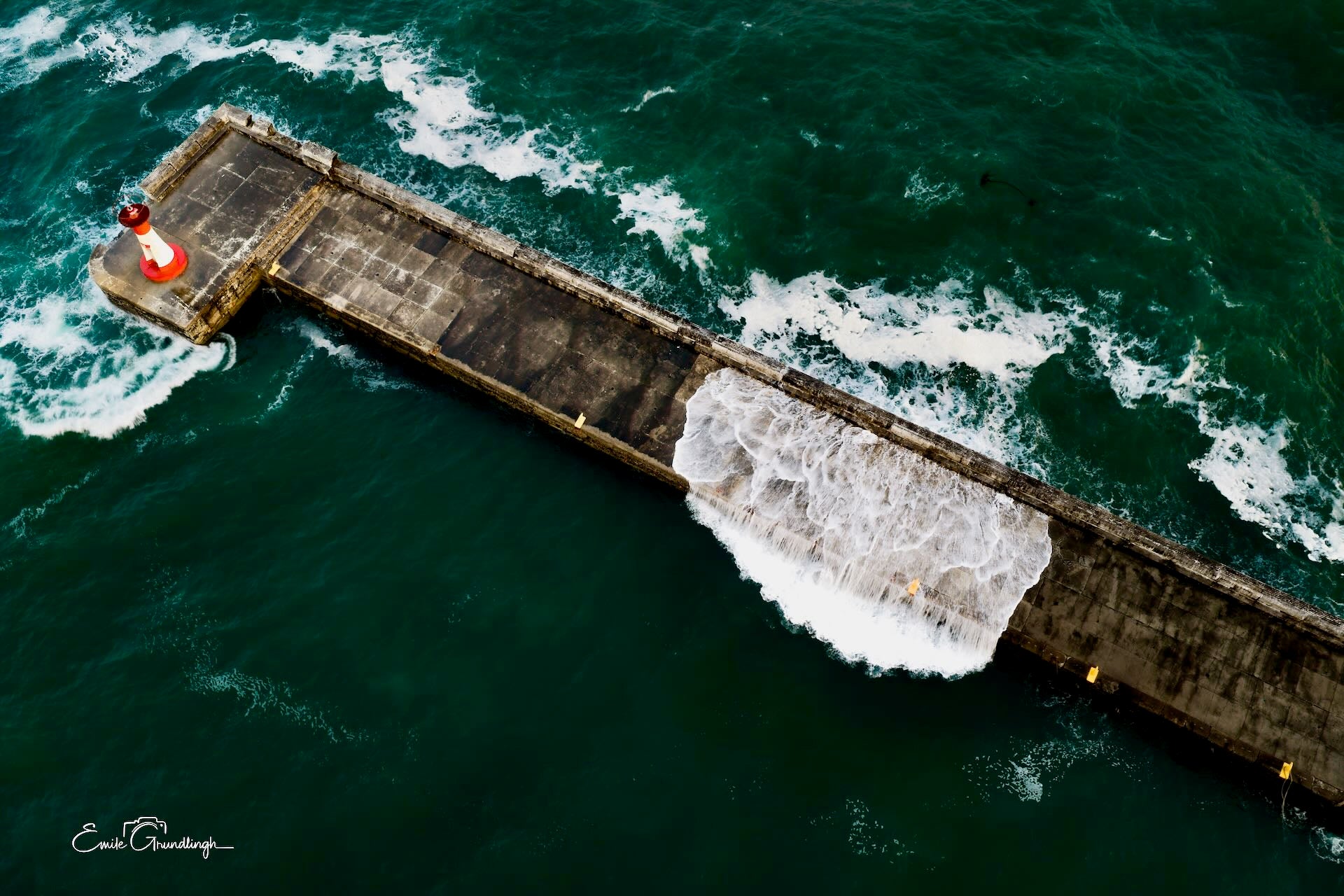 Top-down aerial view of Kalk Bay harbour wall surrounded by emerald green ocean with wave washing across and red lighthouse at end