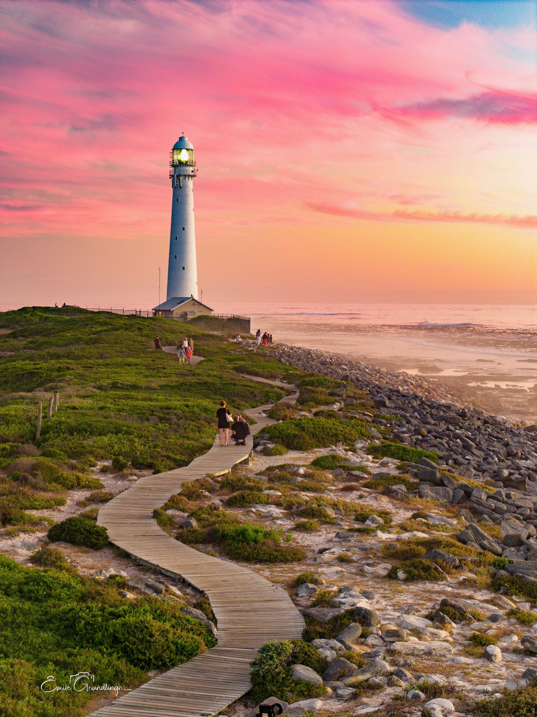 Winding wooden boardwalk leading through coastal fynbos to Slangkop Lighthouse at Kommetjie under a pink and amber sunset sky