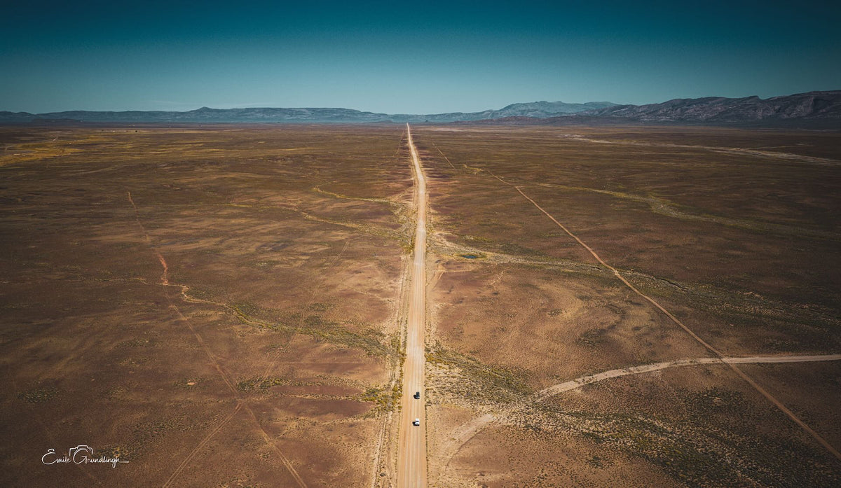 Aerial view of a straight gravel road crossing the vast Tankwa Karoo towards distant mountains beneath a blue sky.