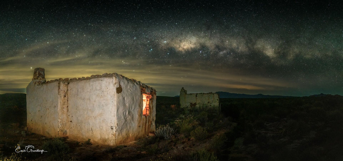 Weathered Karoo house with a warm glowing window beneath the Milky Way, photographed by Emile Grundlingh.