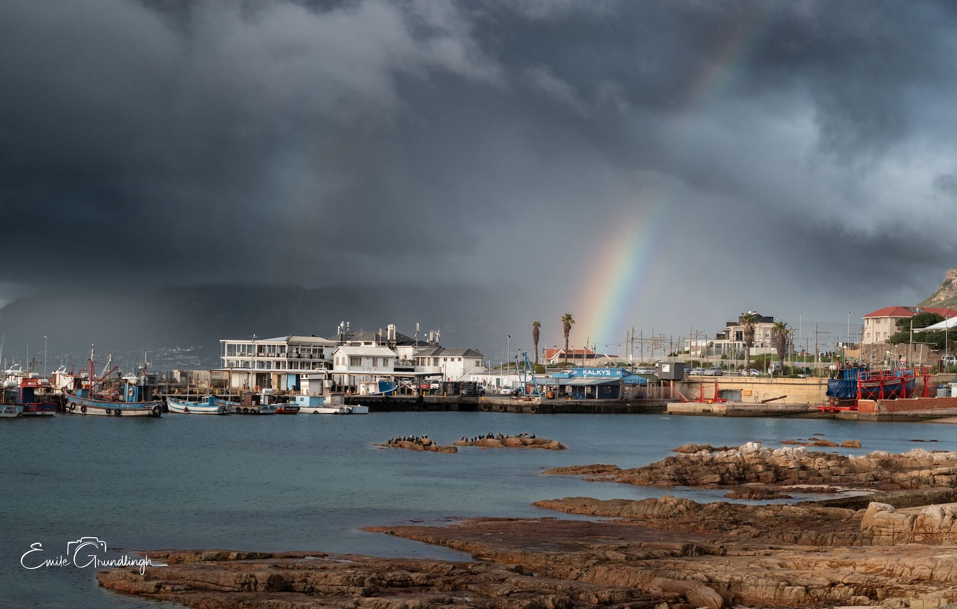 Rainbow arcing over Kalky's fish and chips restaurant at Kalk Bay harbour with fishing boats and cormorants under dramatic storm sky