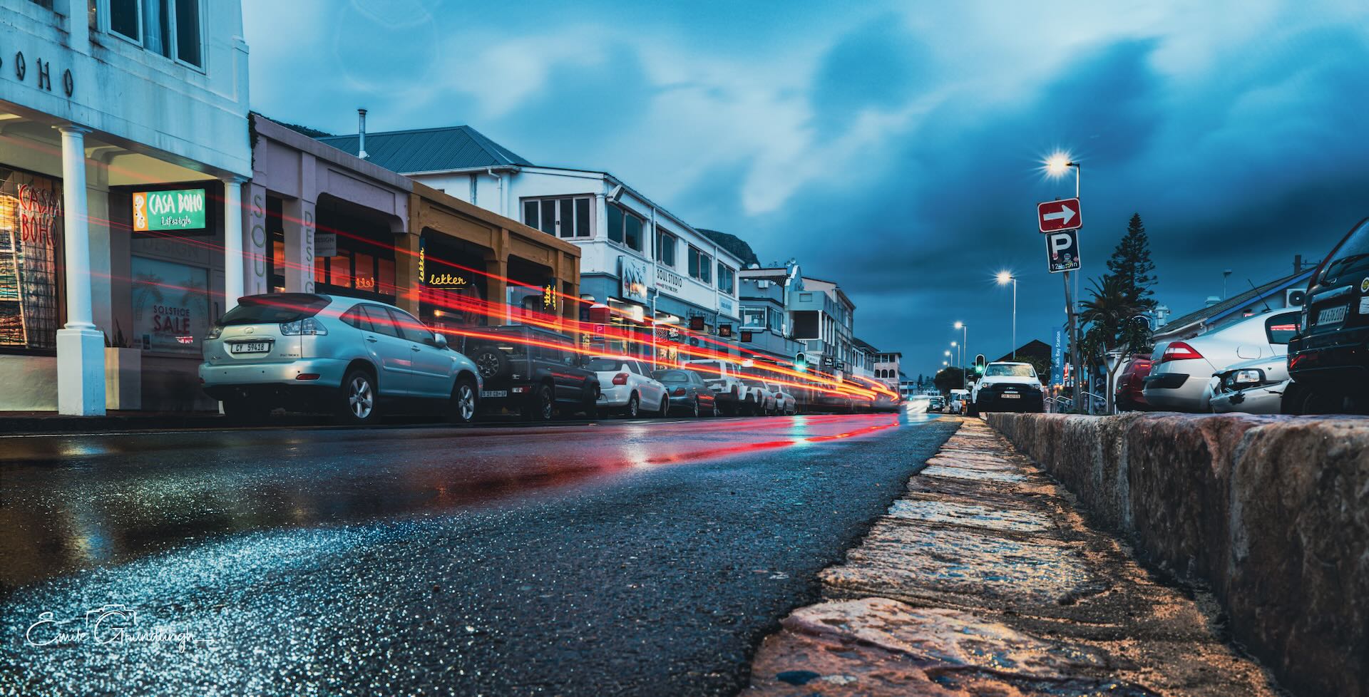 Kalk Bay main road at blue hour with vivid red and orange car light trails reflecting off wet tarmac under moody stormy sky