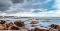 Long exposure seascape of silky water rushing between barnacled boulders at Muizenberg rocky shoreline under dramatic steel blue cloudscape