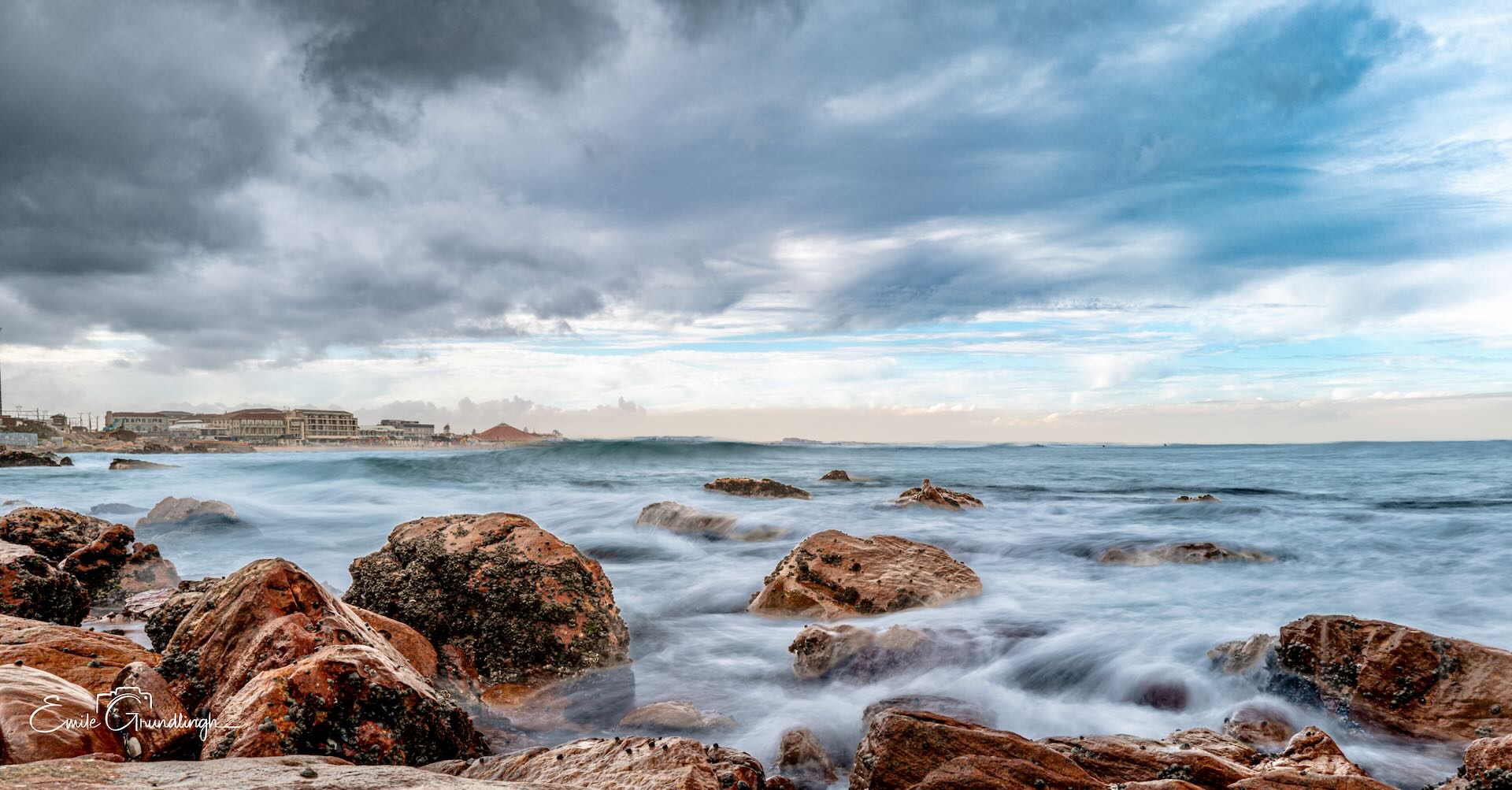 Long exposure seascape of silky water rushing between barnacled boulders at Muizenberg rocky shoreline under dramatic steel blue cloudscape
