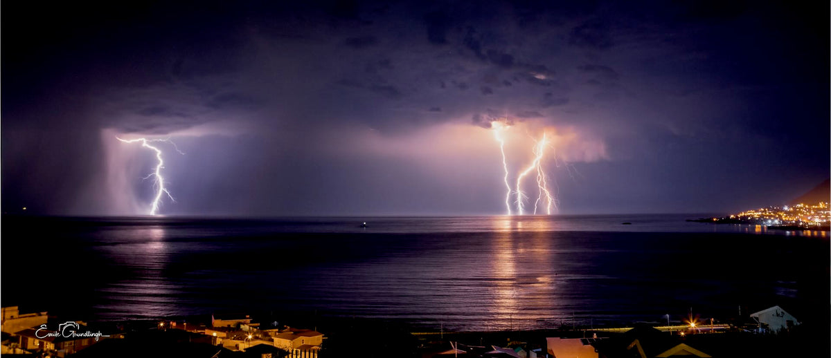 Double lightning strike over a calm ocean bay at night with coastal town lights in the distance