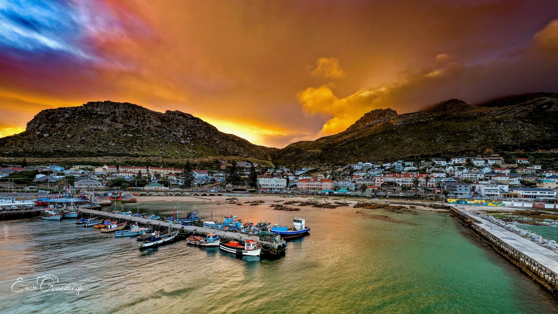 Aerial panorama of Kalk Bay village and harbour under dramatic fiery orange and purple sunset sky with fishing fleet and mountains