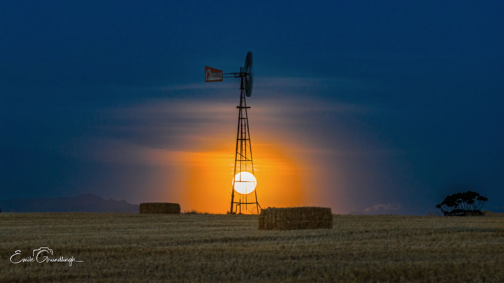 Full moon rising through the silhouette of a windmill above hay bales on the Overberg plains under a dramatic stormy blue sky