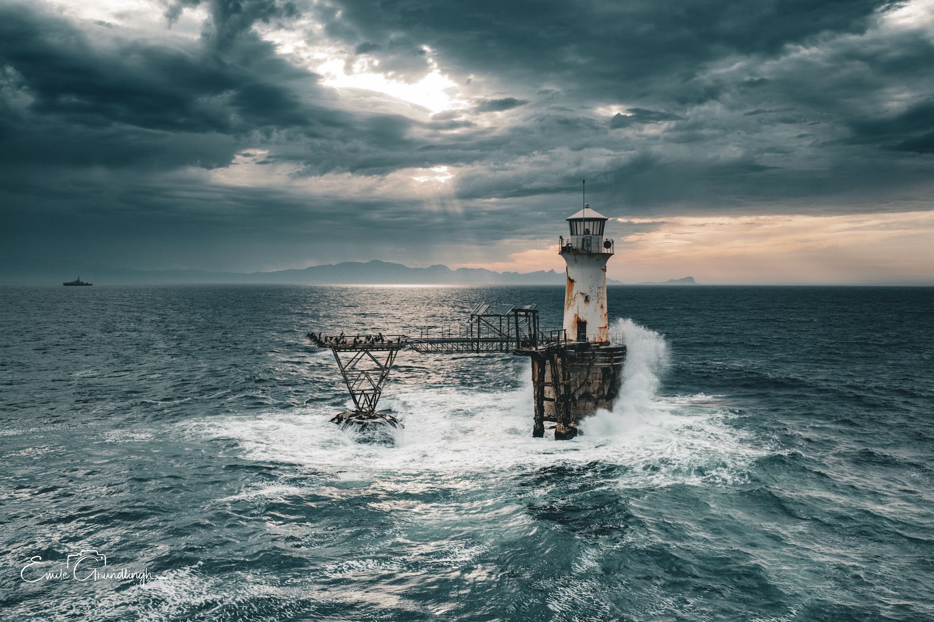 Roman Rock Lighthouse battered by waves with collapsed gantry arm and dramatic god-rays through storm clouds, False Bay