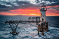 Roman Rock Lighthouse against explosive crimson and magenta sunrise sky with cormorants silhouetted on gantry and mountains on horizon