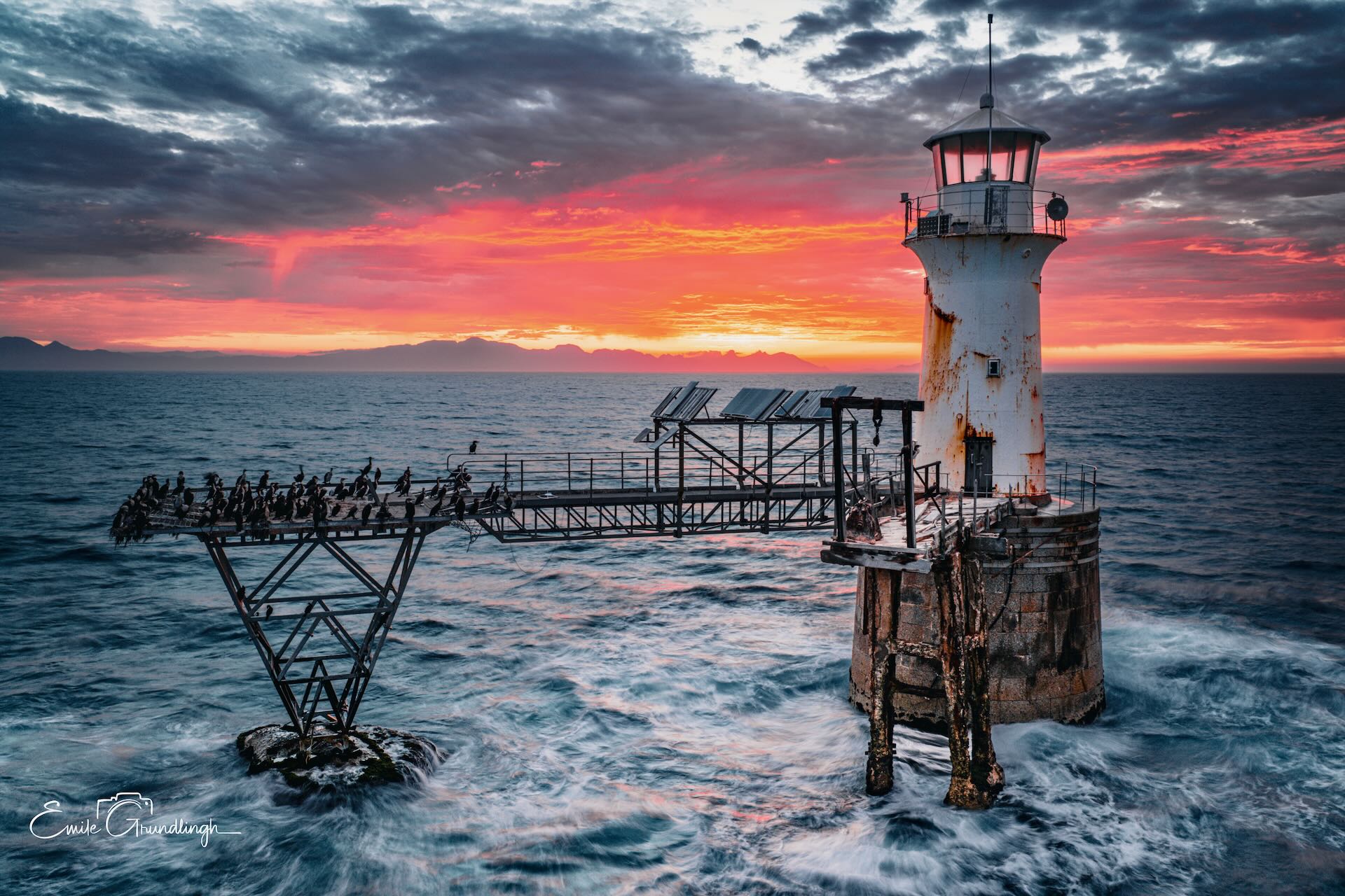 Roman Rock Lighthouse against explosive crimson and magenta sunrise sky with cormorants silhouetted on gantry and mountains on horizon