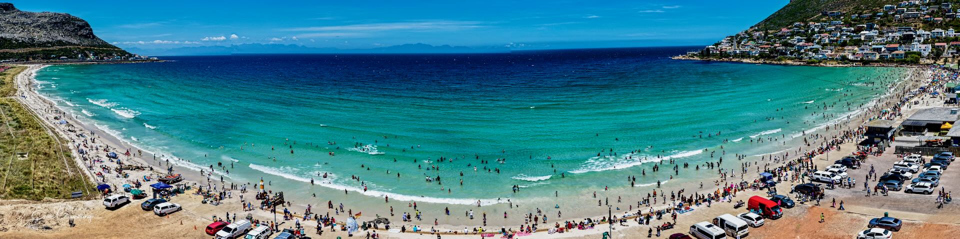 Aerial panorama of Fish Hoek Beach packed with swimmers and sunbathers on New Year's Day with vivid turquoise False Bay waters