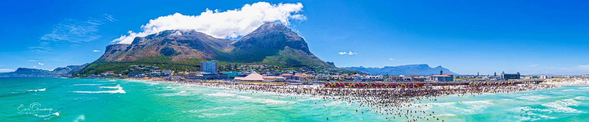 Aerial panorama of thousands of people on Muizenberg Beach on New Year's Day with iconic mountain and pavilion backdrop, False Bay