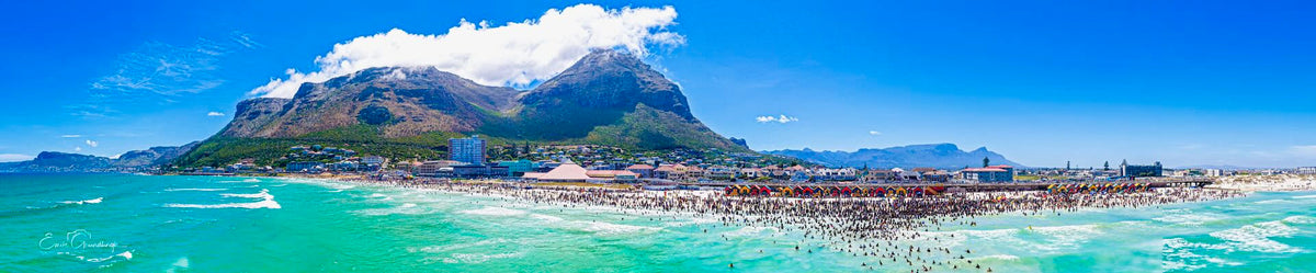 Aerial panoramic view of Muizenberg Beach from the ocean showing turquoise waters, packed summer crowd and Muizenberg Peak in the background