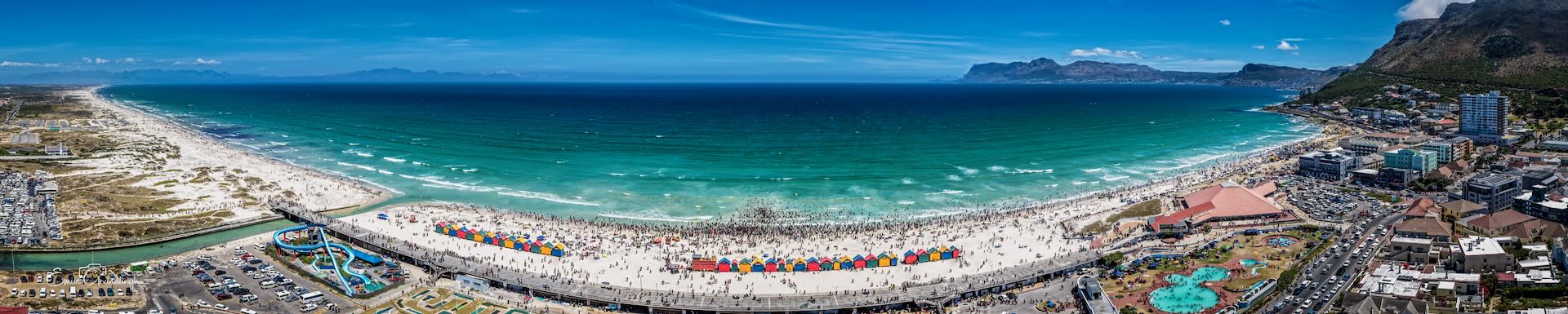 High aerial panorama of Muizenberg Beach showing colourful beach huts, waterpark, crowds and full sweep of False Bay with mountain ranges