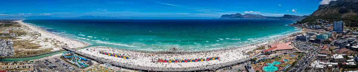 Aerial panoramic view of Muizenberg Beach showing colourful beach huts, crowded summer shoreline, turquoise False Bay waters and mountain backdrop