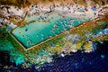 Top-down aerial view of Glen Cairn tidal pool packed with colourful inflatables and swimmers against kelp-covered rocks and deep blue ocean
