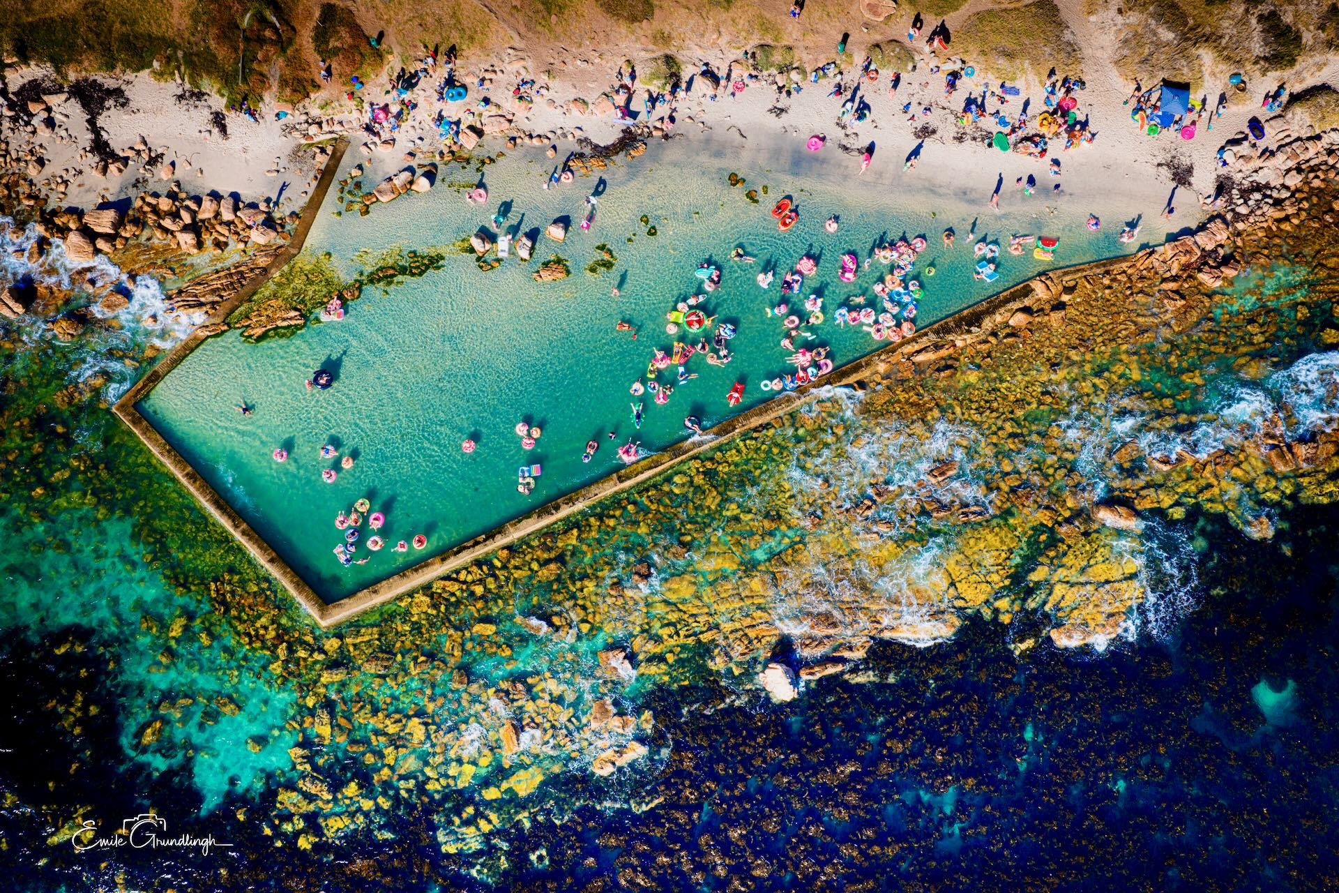 Top-down aerial view of Glen Cairn tidal pool packed with colourful inflatables and swimmers against kelp-covered rocks and deep blue ocean