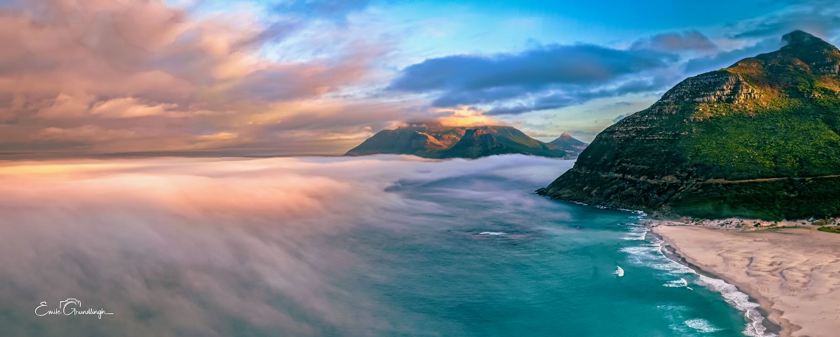 Aerial panorama of sea mist rolling over Noordhoek Long Beach at dawn with Chapman's Peak cliffs lit by golden sunrise light